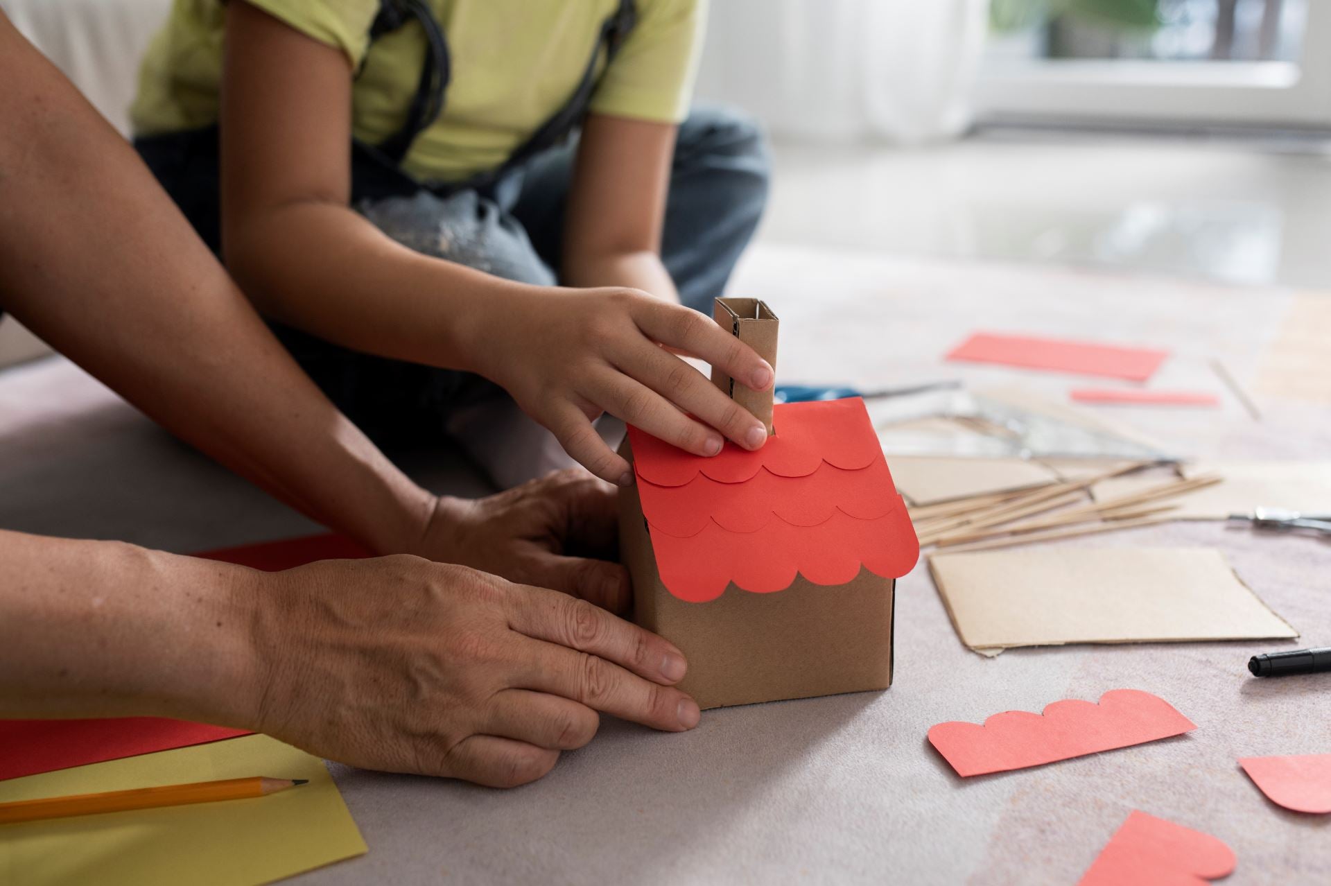 Two people crafting a small cardboard box with red paper roof on a table.