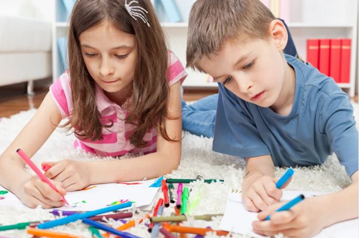 Two children lying on the floor with coloring books and crayons.